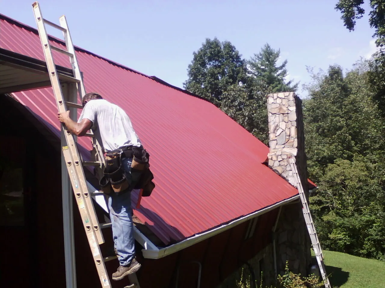 Roofer on a ladder beside a red metal roof and stone chimney — Soffit & Fascia Repair by Roofing Brothers in Adah
